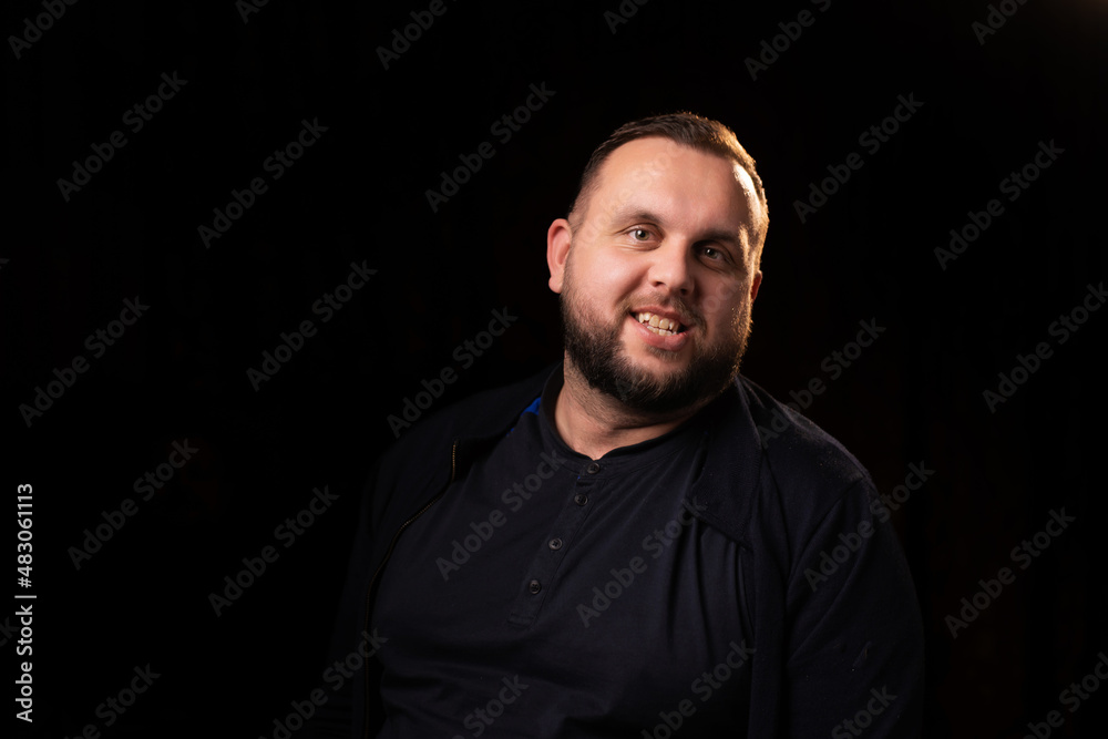 Obraz premium Portrait of a smiling young man with a beard. Dark background. Looks into the camera. Good-natured, contented, happy face.