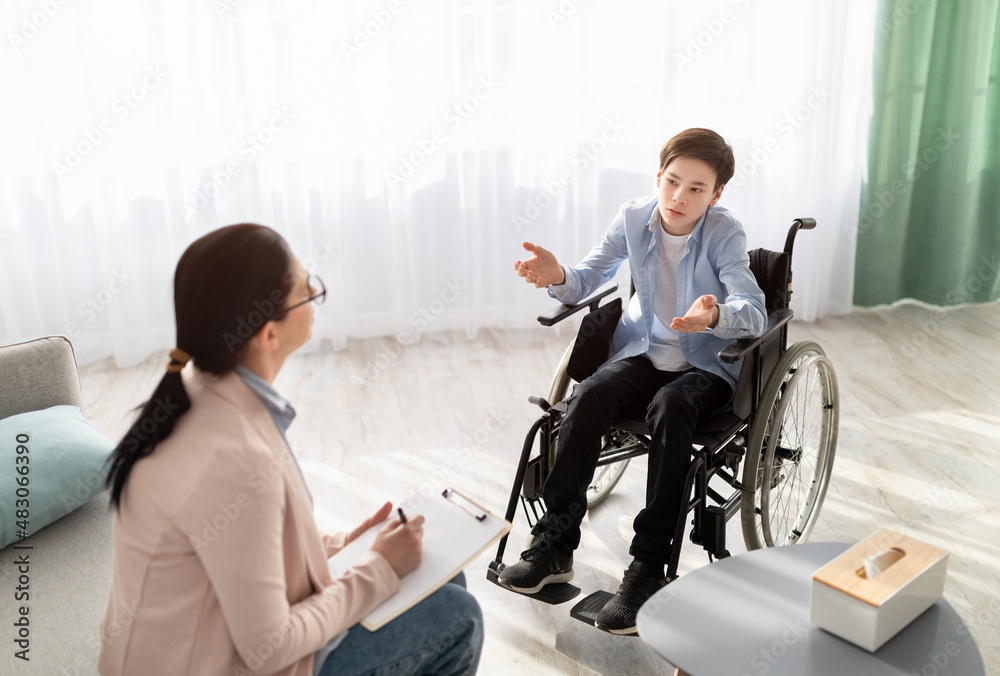 Fototapeta premium Female psychologist consulting disabled teenage boy in wheelchair at office