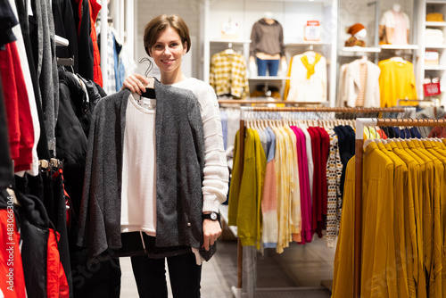 Photo of young attractive brunette woman with a short haircut in a white sweater chooses stylish and casual clothes in a store in a shopping mall. Shop concept