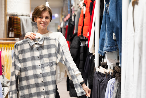 Photo of young attractive brunette woman with a short haircut in a white sweater chooses stylish and casual shirt in a store in a shopping mall. Shop concept