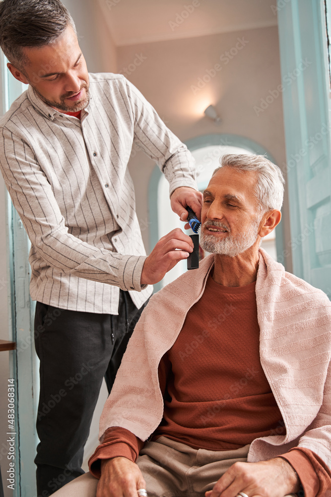 Retirement man looking at his reflection at the mirror while his adult son cutting his beard Retirement man looking at his reflection at the mirror while his adult son cutting his beard