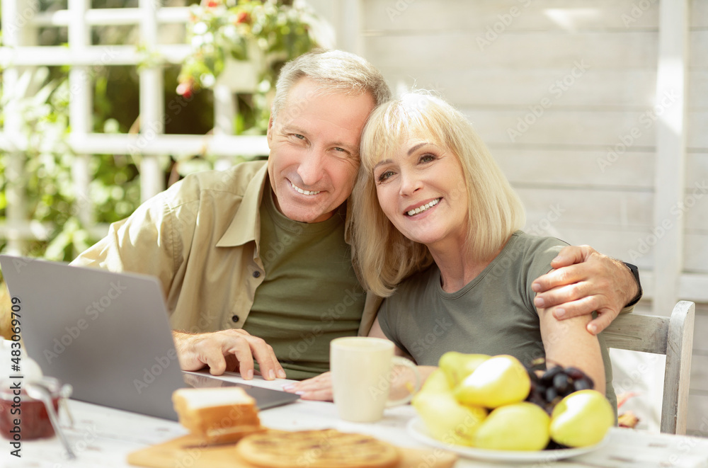 Happy senior couple using laptop computer outdoors, sitting at home terrace and smiling to camera