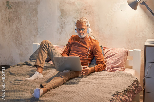 Senior man wearing headphones looking at the laptop while working with it on the bed