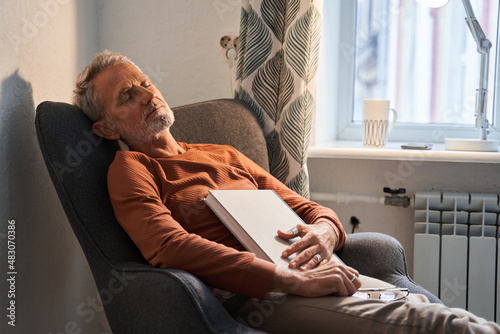 Tired mature man sleeping at the armchair with book on chest after reading photo