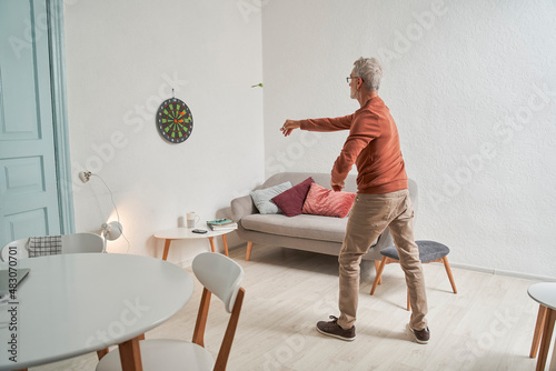 Senior caucasian man throwing darts in cozy room interior, while playing alone at home