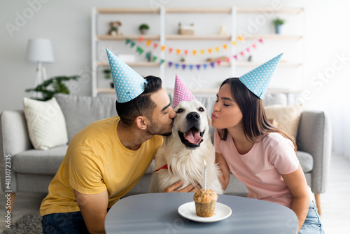 Young interracial couple in party hats celebrating their dog's birthday, kissing their pet at home