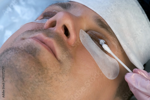 Beautician is applying solution on man's eyelashes for laminating lifting lash procedure. Cosmetologist is making lash lamination in beauty salon, cosmetology clinic, closeup face of client.