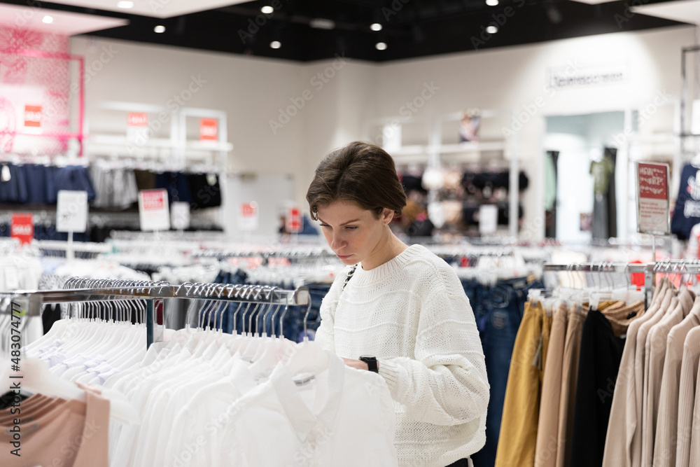 Obraz premium Photo of young beautiful and attractive brunette woman with a short hair in a casual and stylish white sweater chooses fashionable clothes in a store in a shopping mall. Shop interior