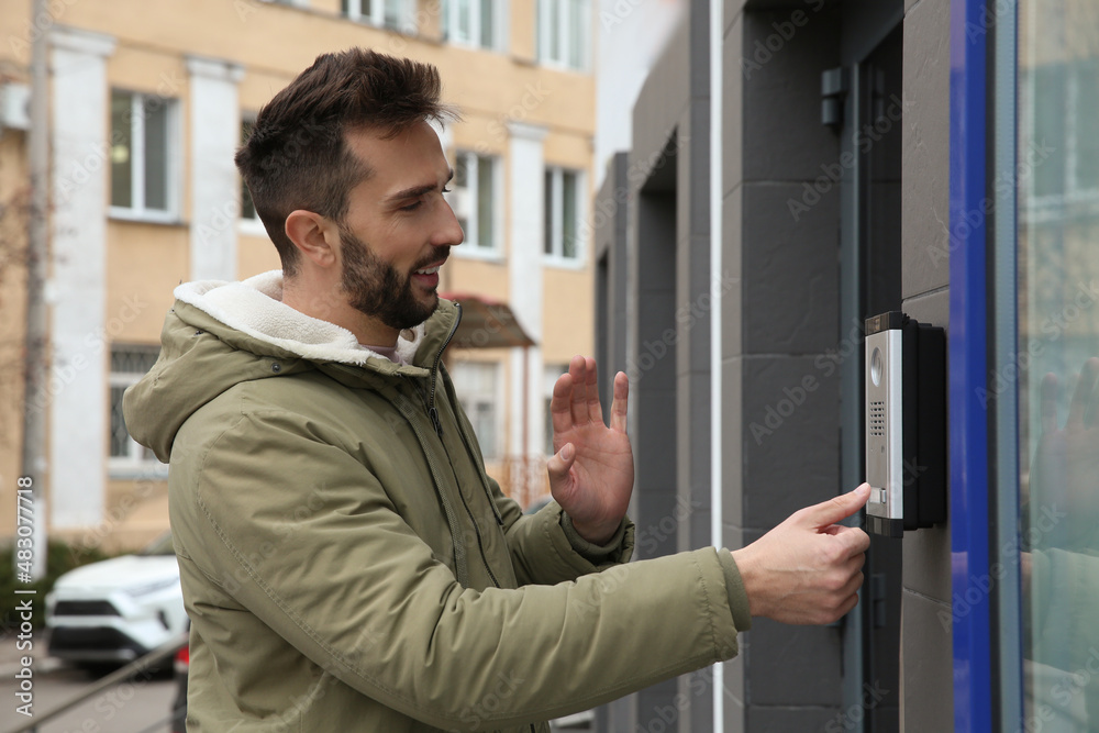Happy man ringing intercom while waving to camera near building entrance Happy man ringing intercom while waving to camera near building entrance
