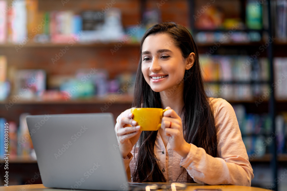 Millennial arab woman enjoying coffee while working on laptop computer in cafe, smiling and looking at pc computer Millennial arab woman enjoying coffee while working on laptop computer in cafe, smiling and looking at pc computer