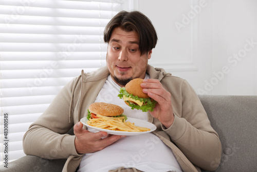 Overweight man with plate of burgers and French fries on sofa at home