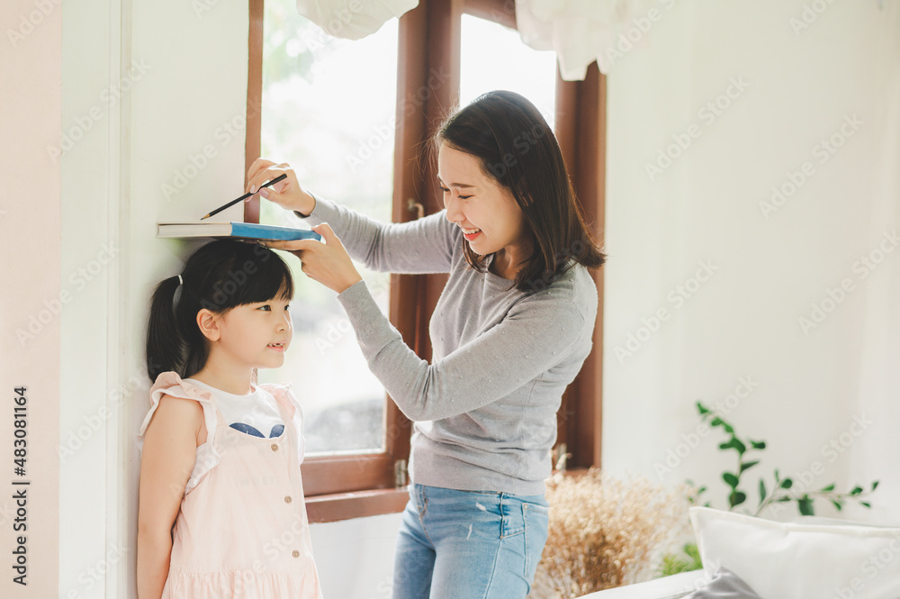 Asian Mother measuring height of her daughter near wall - obrazy, fototapety, plakaty 