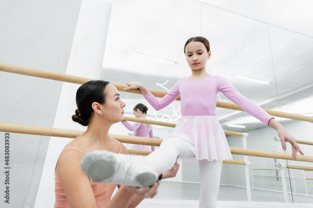 young dance teacher holding leg of girl practicing in ballet school