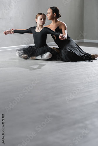 girl in black tutu sitting with crossed legs near ballet teacher in studio photo