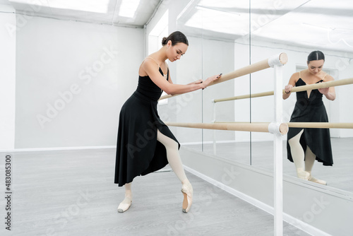 full length view of ballerina in black dress and pointe shoes training at barre near mirrors