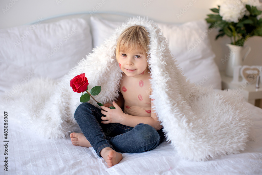 Beautiful blond toddler child, boy with lipstick kisses on his body, holding red rose for Valentine