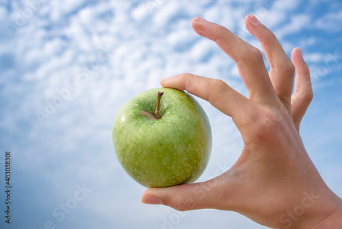 Hand holding green apple with blue sky with clouds as background