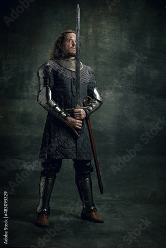 Vintage style portrait of brutal seriuos man, medieval warrior or knight with dirty wounded face holding big sword isolated over dark background. Comparison of eras