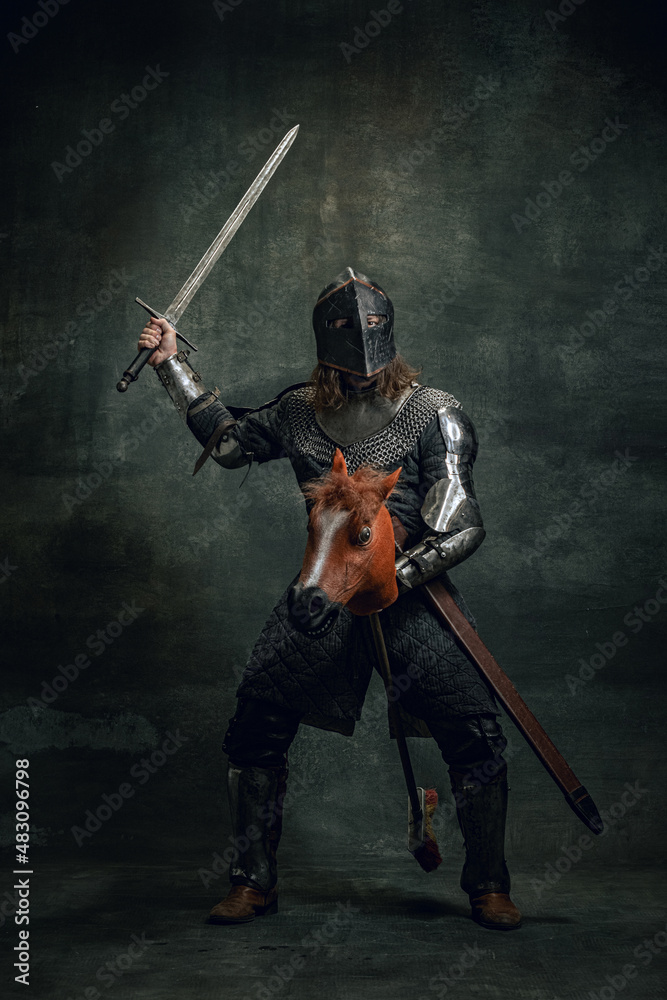 Naklejka premium Portrait of medieval warrior or knight wearing helmet and armor riding toy horse, holding big sword isolated over dark background. Comparison of eras, history