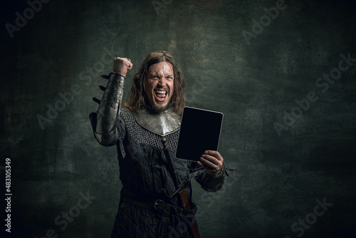 Vintage style portrait of brutal smiling man, medieval warrior or knight with dirty wounded face using modern gadget isolated over dark background. Comparison of eras