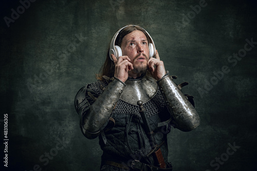 Shocked medieval warrior or knight with dirty wounded face in headphones listening to music isolated over dark vintage background. Comparison of eras, history