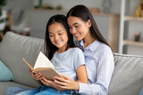 Loving young asian mother reading book with her pretty daughter, sitting together on sofa at home
