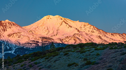Snow covered mountain during sunset with a reddish glow and a blue sky.