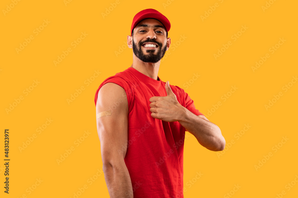 Happy Vaccinated Worker In Red Uniform Gesturing Thumbs-Up, Yellow Background Happy Vaccinated Worker In Red Uniform Gesturing Thumbs-Up, Yellow Background