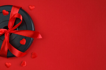 Ribbon wrapped dumbbells weight plate as a love gift for Valentine's Day, birthday, anniversary, wedding. Healthy fitness flat lay composition, gym workout concept, copy space on red background.