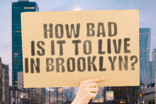 The question " How bad is it to live in Brooklyn? " on a banner in men's hand with blurred background. Danger. Dangerous. Outdoor. Street. Urban. Population. Overpopulation. NYC. New York