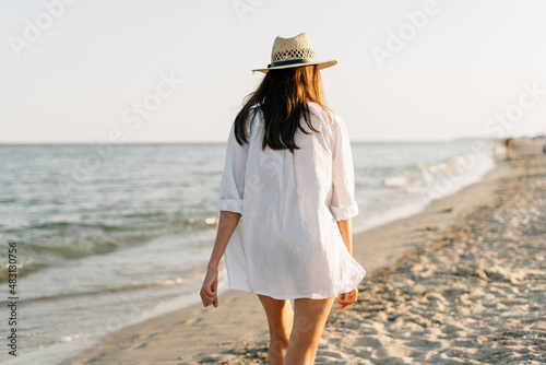 The back of one woman in a shirt and hat walks on a sunny beach with the blue sea in the background. The concept of holidays and vacations.