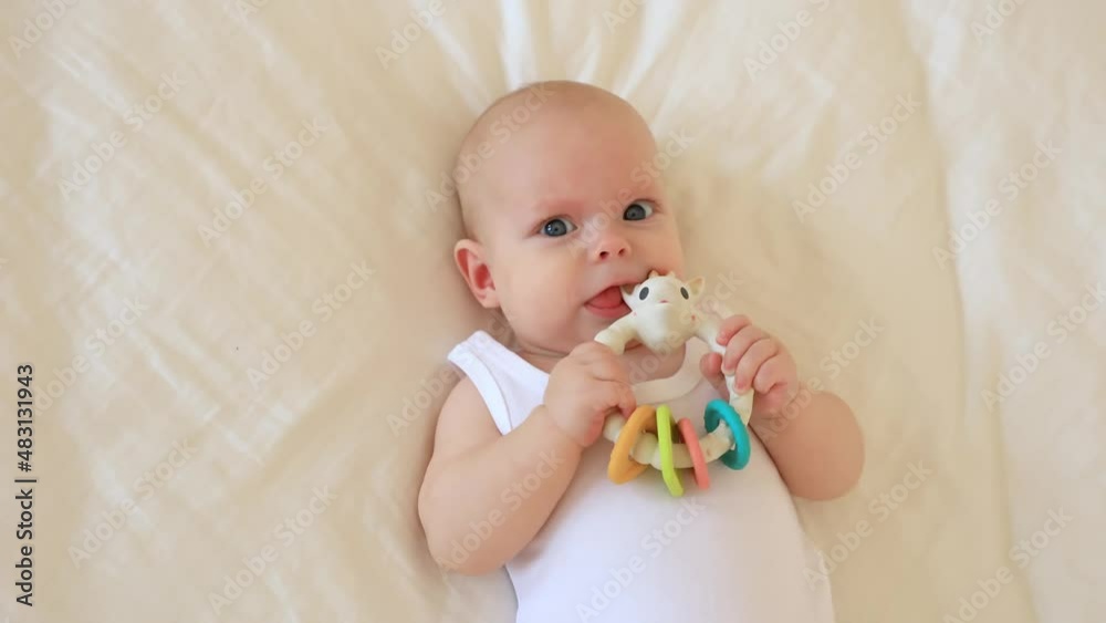 cute baby is lying on his back on a white sheet, holding a rattle, licking, smiling , top view cute baby is lying on his back on a white sheet, holding a rattle, licking, smiling , top view