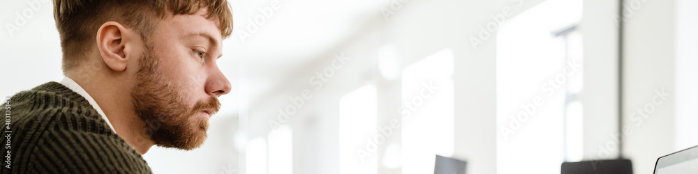 Obraz premium Young ginger man working with laptop while sitting at table in office