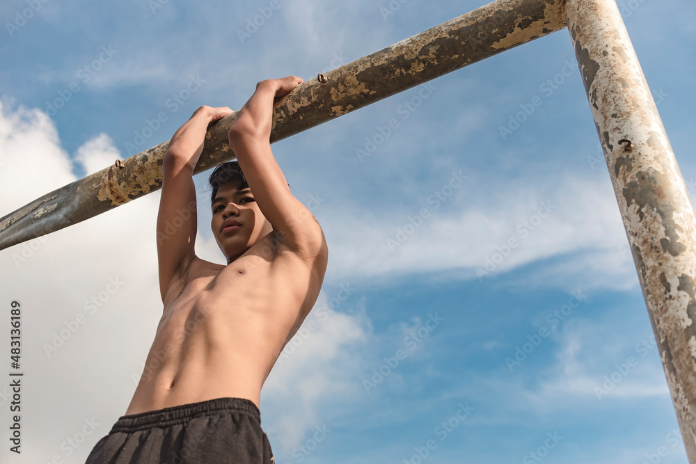 A shirtless teenage man performs chin ups on a thick monkey bar. Upper body and back workout performed outdoors. Stock Photo Adobe Stock