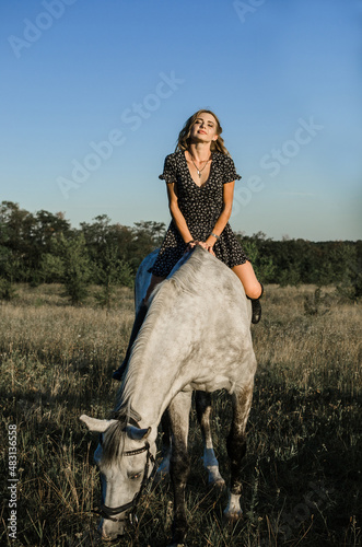 A young beautiful woman sitting astride a white horse with eyes closed in a field in the summer. full-length photo. Rustic, countryside style, Love for animals, pleasure, freedom concept.