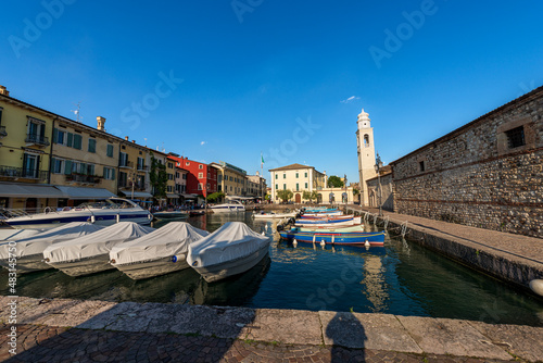 Small port of the Lazise village with moored boats. Tourist resort on the coast of Lake Garda (Lago di Garda). Verona province, Veneto, Italy, Europe. Ancient church of San Nicolo in Romanesque style.