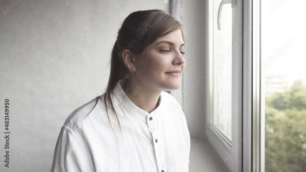 Authentic shot of a pensive young woman while looking through the window and smiling. Concept: life, dreams, hope, new beginning, environment, quarantine, covid-19 Authentic shot of a pensive young woman while looking through the window and smiling. Concept: life, dreams, hope, new beginning, environment, quarantine, covid-19