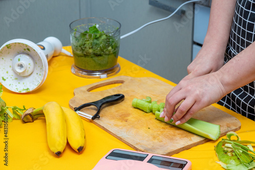 Women's hands cut celery on wooden board with knife. There are bananas nearby, blender bowl with greens. Selective focus.