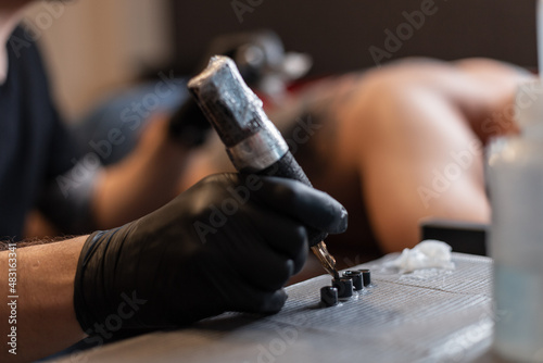 Male hand in a black glove with a tattoo machine takes ink and makes a tattoo in the studio. Man professional tattoo artist in work process, close-up photo