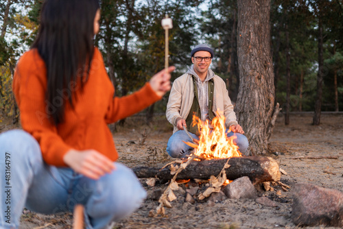 Look at this. Full length view of the caucasian woman showing stick with marshmallow to her husband while sitting around the fire and frying. People talking with each other. Forest at the background
