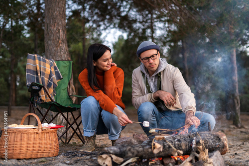 Full length view of the man and woman sitting around the fire and frying marshmallows. People talking with each other. Forest at the background