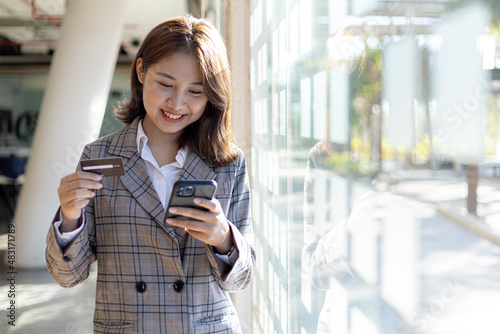 A woman standing by a window holding a cell phone and credit card, she is shopping online on a smartphone, she pays for goods and services by credit card. Concept of using credit card for payment.