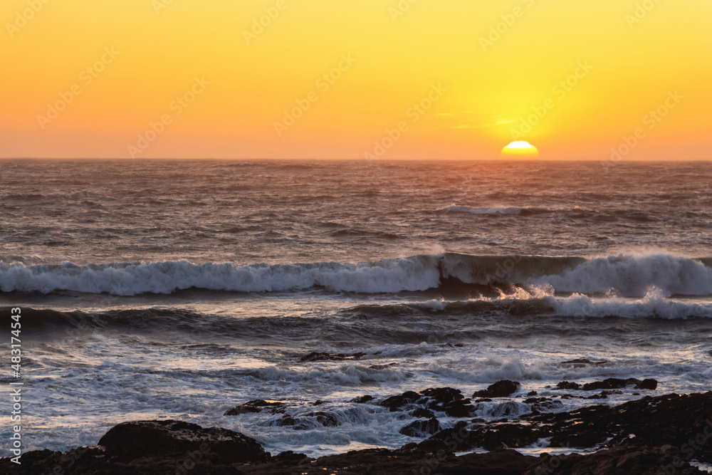 A picturesque orange sunset over the Atlantic Ocean, with the sun halfway below the horizon and foaming waves crashing against the dark rocks on the shore