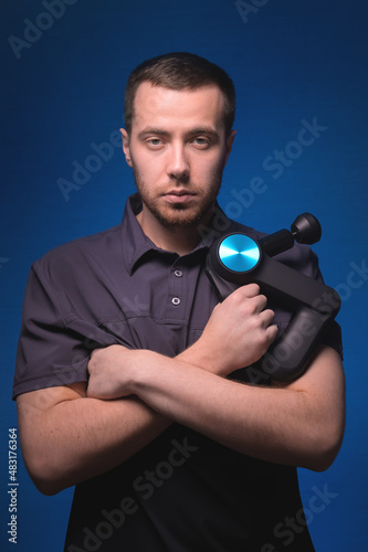 Portrait of a confident professional masseur with a percussion massager in his hands. Low key, blue background. Shock wave massage
