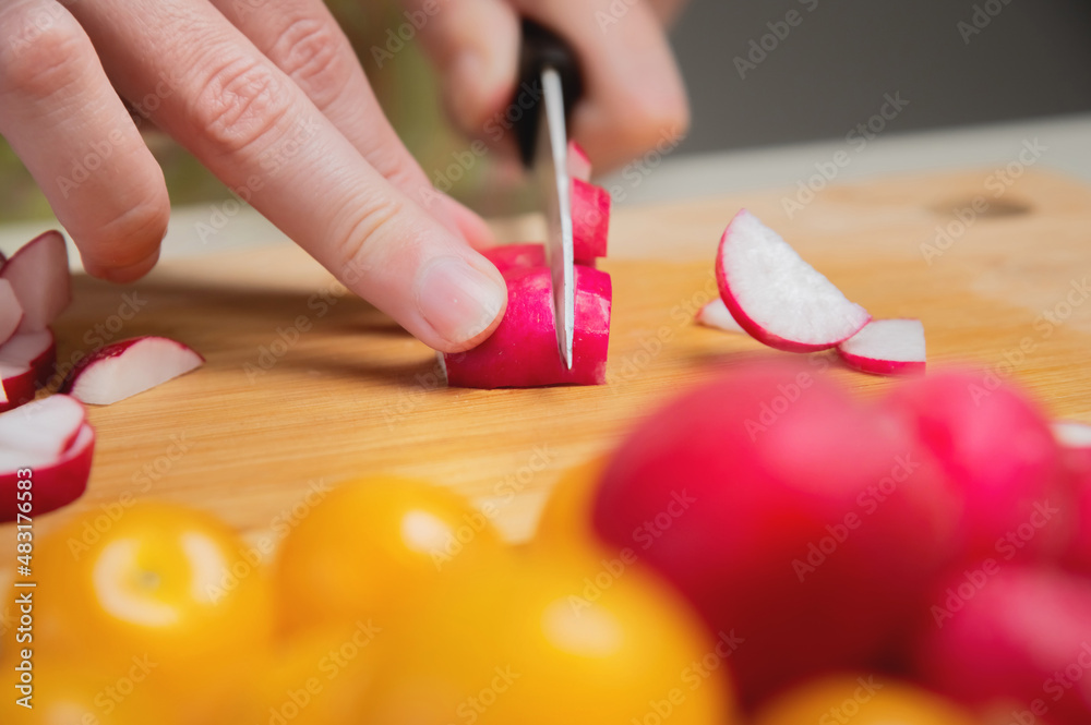 Close-up of female hands cut fresh raw radish on a wooden board against the background of yellow cherry tomatoes. Vegetarian tasty and healthy food Close-up of female hands cut fresh raw radish on a wooden board against the background of yellow cherry tomatoes. Vegetarian tasty and healthy food