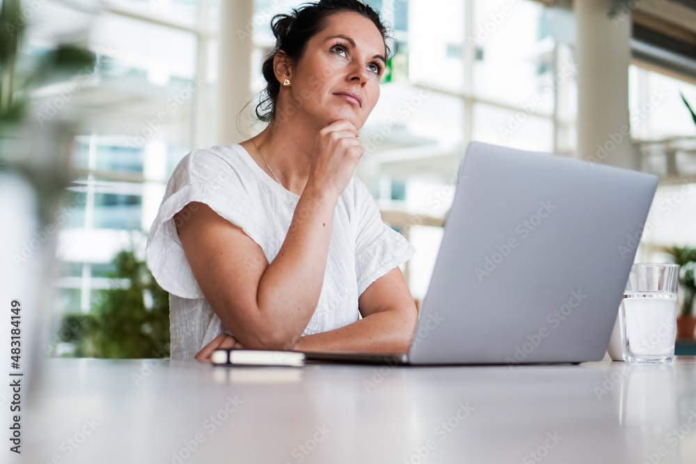 Fototapeta premium thoughtful brooding remote working dark haired woman sitting infront of a laptop or notebook in casual outfit on her work desk in her modern airy bright living room home office with many windows 