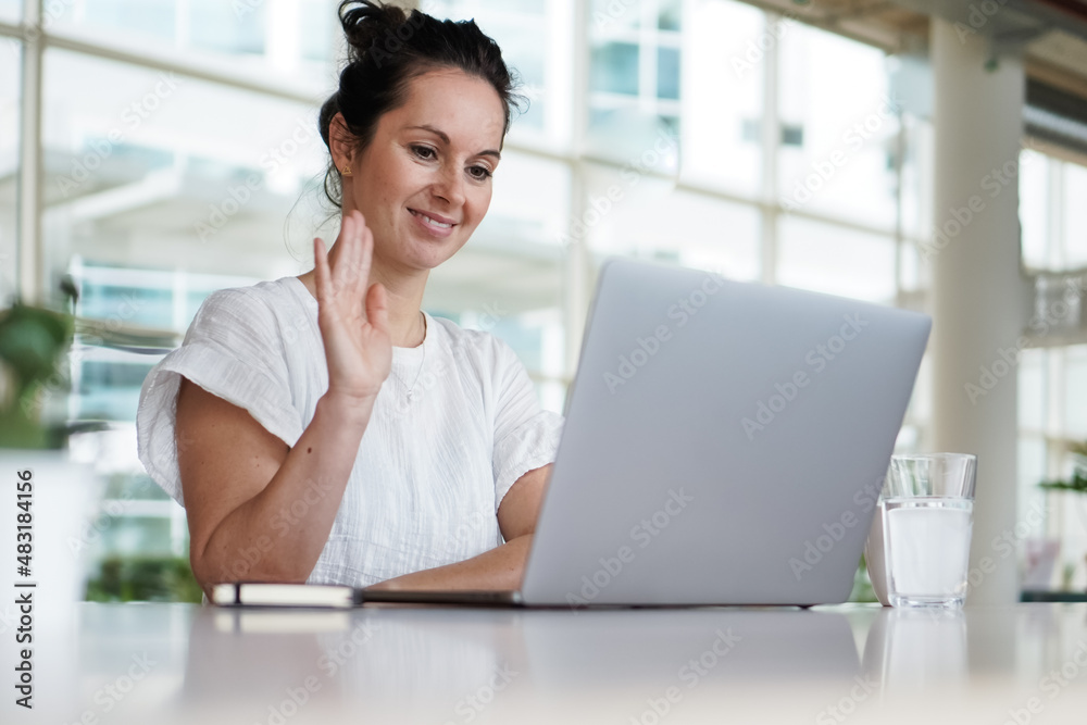 smiling and greeting remote working woman waving hand to laptop or notebook in casual outfit sitting on her work desk in her modern airy living room home office while having a video chat conference smiling and greeting remote working woman waving hand to laptop or notebook in casual outfit sitting on her work desk in her modern airy living room home office while having a video chat conference