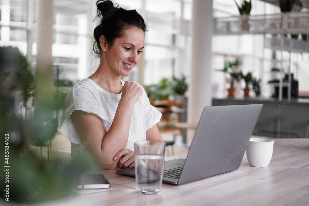 happy smiling remote working woman talking to laptop or notebook in casual outfit sitting on her work desk in her modern loft living room home office having a video chat