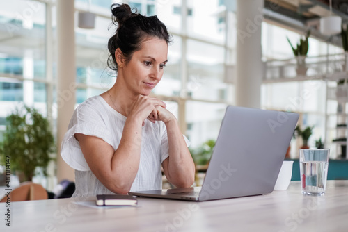 thoughtful brooding remote working dark haired woman sitting infront of a laptop or notebook in casual outfit on her work desk in her modern airy bright living room home office with many windows 