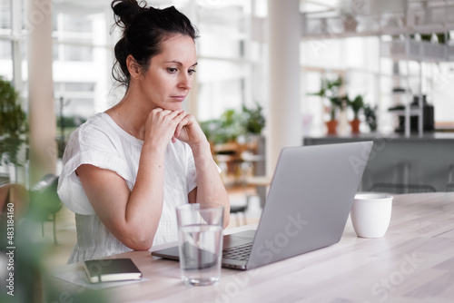 thoughtful brooding remote working dark haired woman sitting infront of a laptop or notebook in casual outfit on her work desk in her modern airy bright living room home office with many windows 